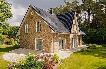 ECO System HAUS – Friesenhaus mit rot-braunem Stein, weißen Fenstern und dunklem Dach im Grünen