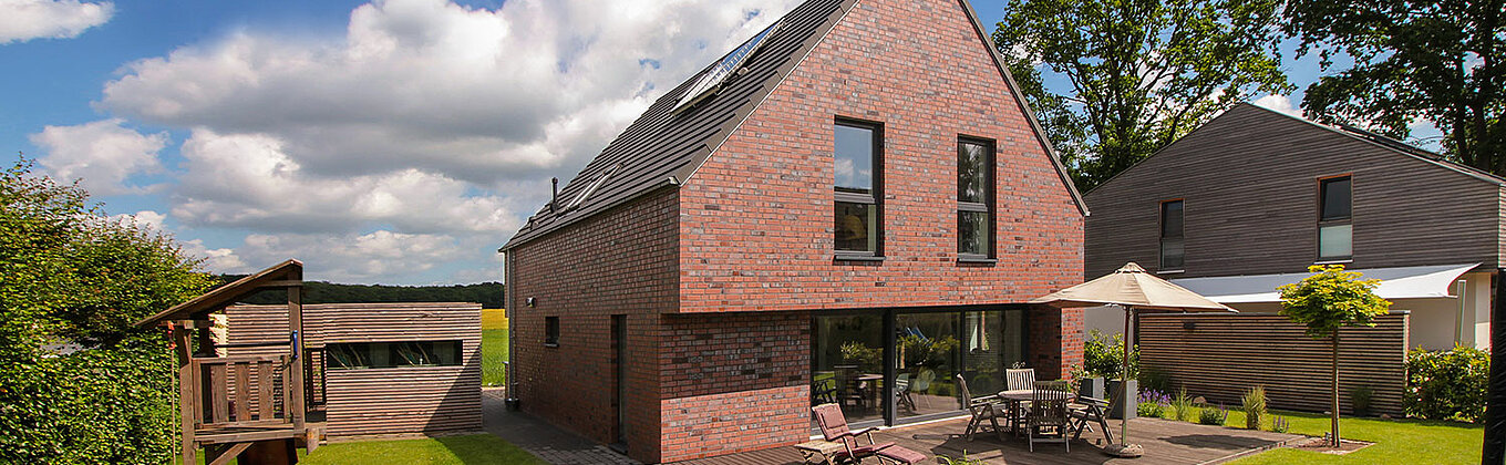 ECO System HAUS – Vario-Haus mit rotem Stein, dunklem Dach und dunklen Fenstern, Terrasse und Garten