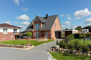 ECO System HAUS – Kapitänshaus mit rotem Stein, dunklem Dach, weißen Fenstern und Vorgarten und einer Garage